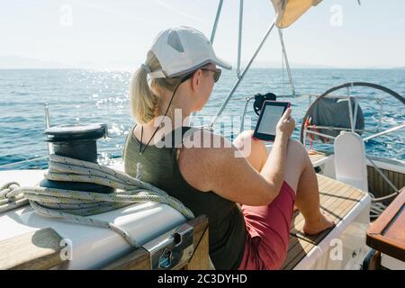 Frau, die an Deck einer Yacht sitzt und dabei genießt, in ihrem eBook zu lesen. Reise- und Urlaubskonzept. Stockfoto