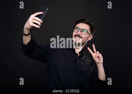 Lächelnder, hübscher junger Mann in einer Brille, der ein Friedenszeichen zeigt, während er Selfie auf dem Smartphone nimmt Stockfoto
