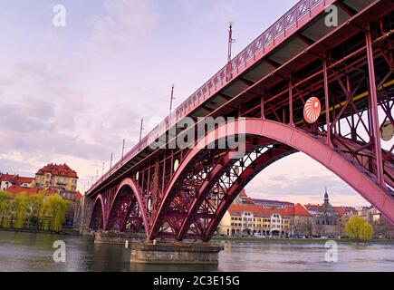 Maribor, Slowenien, Europa. Hauptbrücke (Glavni most, Stari most) über die Drau. Beliebte Flussufer Fastenzeit im Hintergrund. Sonnenuntergang und Abend. Stockfoto