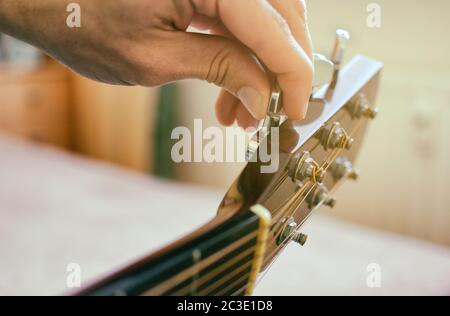 Tuning der Gitarre. Finger drehen den Stimmwirbel auf dem Kopf der akustischen Gitarre. Authentische Aufnahme mit unscharfem Raum im Hintergrund. Stockfoto