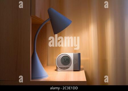 Detail der gemütlichen Schlafzimmer - Leselampe und Wecker auf Nachttisch aus Holz. Einfarbiger Vorhang im Hintergrund. Stockfoto