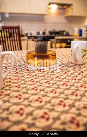 Glaspudding mit Tee und Tassen auf dem Küchentisch im Hintergrund der Küche. Für Text platzieren. Das Konzept des glücklichen Familienlebens Stockfoto