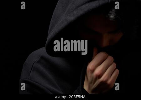 Ein junger Mann in einem schwarzen Hoodie auf schwarzem Hintergrund mit seinem Gesicht bedeckt. Stockfoto