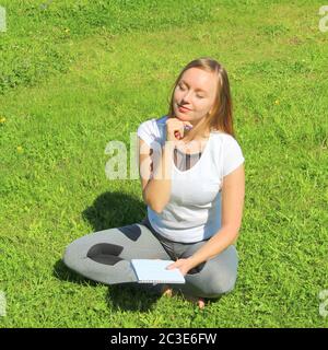 Ein junges, schönes Mädchen im weißen T-Shirt mit geschlossenen Augen sitzt auf dem grünen Gras auf dem Rasen mit einem Notizbuch und einem Stift, mit dem Handkinn gestützt, denkt und träumt, komponiert Text und Gedichte. Stockfoto