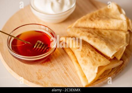 Zwei kleine Glasschüsseln mit Sauerrahm und frischem Honig stehen auf dem Schneidebrett mit drei zusammengefalteten hausgemachten Pfannkuchen Stockfoto