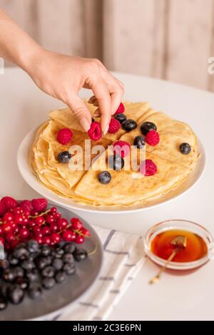 Hand der jungen Frau oder Hausfrau, die frische reife Himbeere von oben auf hausgemachte Pfannkuchen auf dem Teller beim Frühstück Stockfoto