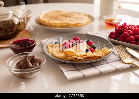 Frisch hausgemachte appetitliche Crepes mit Beeren und Honig auf dem Teller, Schüsseln mit Kirschmarmelade und Schokoladencreme, Teekanne und reifen Himbeeren Stockfoto