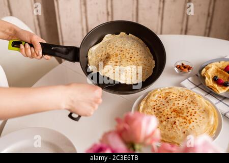 Hände der jungen Frau halten Bratpfanne über Küchentisch, während heiße appetitliche Pfannkuchen, um es auf andere Crepes auf dem Teller setzen Stockfoto