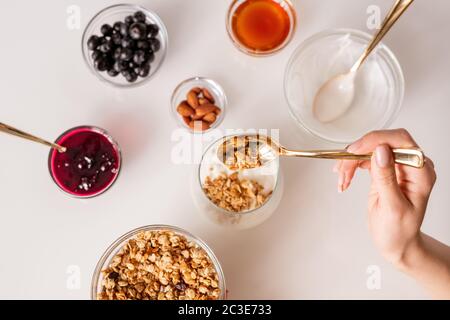 Hand der jungen Frau mit Teelöffel setzen Müsli in Glas mit frischer Sauerrahm während der Herstellung Joghurt mit Marmelade, Mandelnüsse, Honig und Beeren Stockfoto