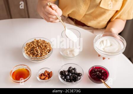 Junge Frau macht hausgemachten Joghurt mit Sauerrahm, Müsli, Honig, Mandelnüsse, frische Brombeeren und Kirschmarmelade zum Frühstück Stockfoto