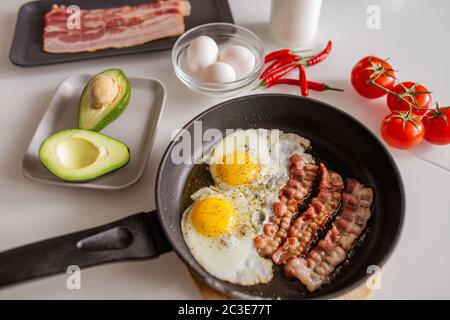 Bratpfanne mit Spiegeleiern und Speck bestreut mit Gewürzen, frischer Avocado, roten reifen Tomaten und scharfem Chili auf dem servierten Küchentisch Stockfoto