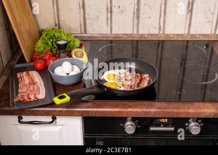 Bratpfanne mit Spiegeleiern und Speck auf Elektroherd mit frischen reifen Tomaten, Avocado und Salat in der Nähe in der Küche Stockfoto