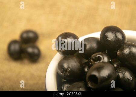 Schwarze kernlose Oliven in einer Keramikschale und ein paar verstreute Oliven auf einem Hintergrund aus grobstrukturiertem Gewebe. Nahaufnahme Stockfoto