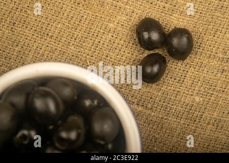 Schwarze kernlose Oliven in einer Keramikschale und ein paar verstreute Oliven auf einem Hintergrund aus grobstrukturiertem Gewebe. Nahaufnahme Stockfoto