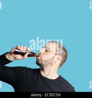 Dunkelhaariger bärtiger Mann in dunkler Kleidung trinkt mit geschlossenen Augen ein dunkles Getränk aus einer Flasche Stockfoto