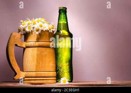 Bierkrug aus Holz mit feinen Gänseblümchen ähnlich Schaum und einer grünen vollen Flasche mit einem Metalldeckel Stockfoto