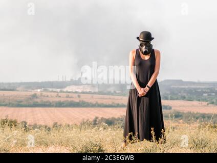 Junge Mädchen in einem schwarzen Kleid und Gasmaske auf dem Hintergrund des rauchenden Fabrikschloten in der Ukraine Stockfoto