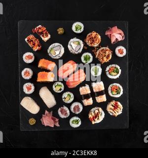 Großes Sushi-Set, von oben auf quadratischem Schiefer auf schwarzem Hintergrund geschossen. Eine Auswahl an verschiedenen Maki, Nigiri und Brötchen Stockfoto