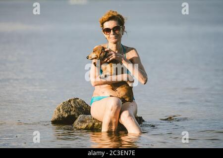 Reife kaukasische Frau sitzt im Wasser auf Stein und halten Hund von Dachshund Rasse in Armen. Thema Hitze und Sommerurlaub. Strandurlaub am Fluss mit Stockfoto