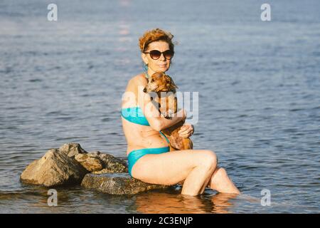 Reife Frau genießen Tag am Strand mit Hund. Die Besitzerin, Hündin im Badeanzug und kleiner, brauner Dackel-Hund, verbringt ihre Sommerzeit in Kälte Stockfoto