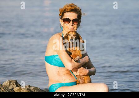 Ein kleiner brauner, alter Hund einer Dachshund-Rasse sitzt auf den Händen einer reifen Frau in einem Teich. Thema, um den Sommer Schwimmen im Fluss mit einem Haustier zu verbringen Stockfoto