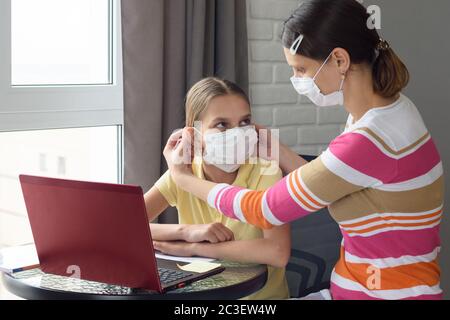 Mama legt einem Mädchen eine medizinische Gesichtsmaske zu, während sie zu Hause vor einem Computer sitzt Stockfoto