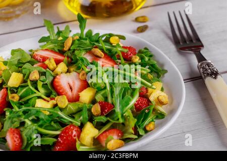Frischer Salat mit Rucola, Erdbeeren, Ananas und Pistazien auf weißem Teller auf rustikalem Holztisch. Organisches Konzept. Stockfoto