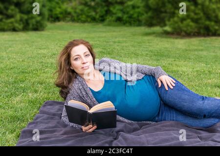 Schwangere Frau liest ein Buch auf einem grünen Feld in einem Park. Stockfoto