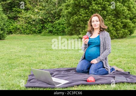 Schöne schwangere Frau lächelt und entspannt in einem Park mit Äpfeln und Laptop. Stockfoto