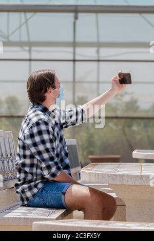 Junger Mann mit einer OP-Maske sitzt an einem Tisch und macht ein Foto von sich selbst mit seinem Telefon auf einem unscharf Hintergrund. Sicherheit und neues Leben Stockfoto