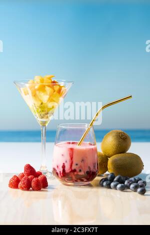Köstlicher Smoothie mit einem Stroh, umgeben von Heidelbeeren, Kiwis, Himbeeren und einem martiniglas, gefüllt mit Aprikose, Ananas und Kiwi-Stückchen auf einem O Stockfoto