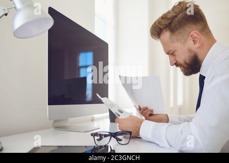 Ein ernstzunehmender Unternehmer liest Arbeitsdokumente, die mit einem Computer im Büro an einem Tisch sitzen. Stockfoto