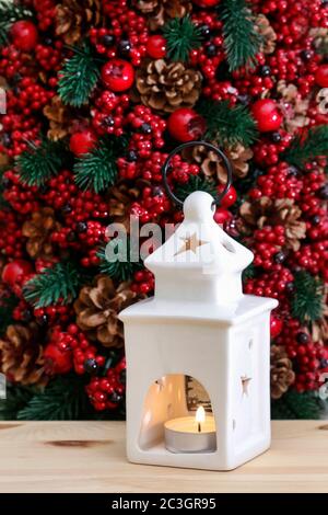 Weiße Keramik Laterne mit einer Kerze auf rotem Weihnachtshintergrund aus Tanne, Beeren und Zapfen. Festliche Einrichtung Stockfoto