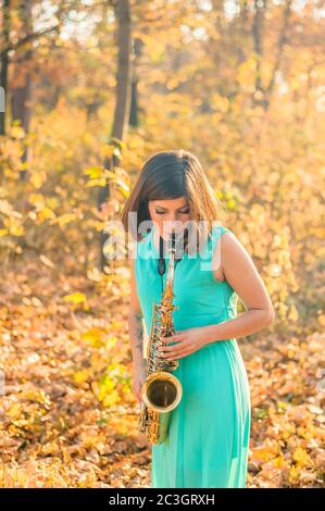 Schönes junges Mädchen mit schwarzen Haaren und einem Tattoo am Arm, in einem langen blauen Kleid gekleidet und spielt im Herbst das Altsaxophon Stockfoto