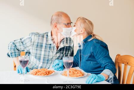 Bild eines älteren Ehepaares, das beim Abendessen mit Masken küsst und blaue Latexhandschuhe trägt - Lebenskonzept mit COVID (Coronavirus), romantische Nutzung der Pers Stockfoto