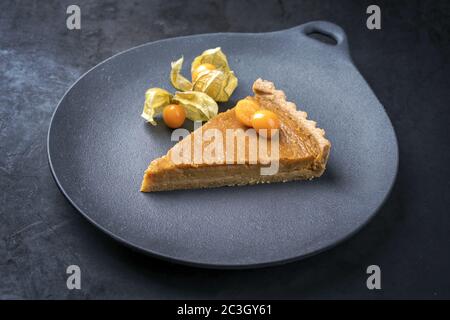 Traditionelles französisches Stück Tarte au Citron mit Orange und Physalis-Frucht, angeboten als Nahaufnahme auf einem modernen Design-Teller Stockfoto