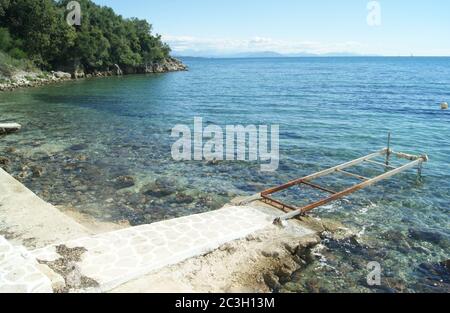 Ein alter Metallsteg an der Küste auf der schönen griechischen Insel Korfu. Bewaldete Klippen am Wasser Rand in der ruhigen, Familien, Resort von Nissaki auf t Stockfoto