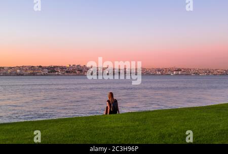 Tourist Girl mit Rucksack sitzt auf einem Gras im Jardim do Rio Park am Ufer des Flusses Tejo und genießt den Sonnenuntergang über Lissabon. Stockfoto