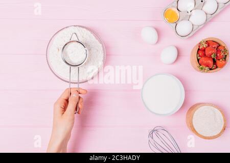 Prozess der Herstellung von Teig für Erdbeerkuchen, weibliche Hand sieben das Mehl neben den Zutaten, Eier, Zucker und frische Beeren auf einem rosa Tisch, Top Stockfoto