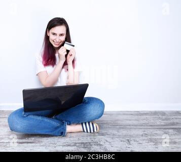 Junge Frau auf dem Boden sitzend Laptop hält glücklich lächelt und in den Händen Kreditkarte, grauer Hintergrund. Kopierbereich Stockfoto