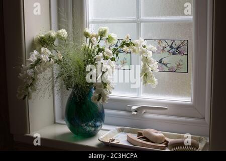 Weiße Blumen und Laub in einer grünen Glasvase auf einer Fensterbank Stockfoto