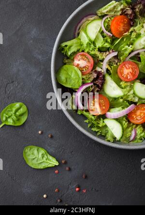 Frischer, gesunder vegetarischer Gemüsesalat mit Tomaten und Gurken, roter Zwiebel und Spinat in großer, grauer Schüssel auf dunklem Hintergrund. Draufsicht. Mac Stockfoto