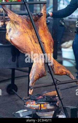 Mann, der eine ganze Fleischkadaver auf einem Spieß über einem kleinen Feuer in einem Brazier im Freien bei einer Veranstaltung oder Party in der Nähe röstet Stockfoto