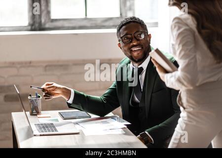 Serious junger afrikanischer Mann arbeitet mit jungen schönen Frau an seinem modernen Coworking Platz mit Laptop zusammen Stockfoto