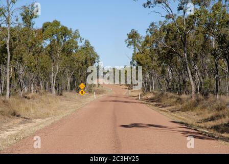 Versiegelte Straße durch Eukalyptusbäume im Outback mit Straßenverengen und Warnschildern für Flutwege, in der Nähe von Charters Towers, Queensland, Australien Stockfoto