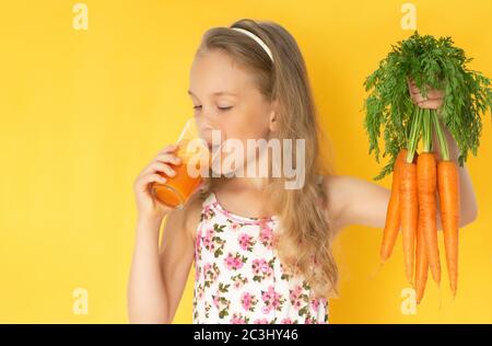 Kleines Mädchen, das auf ein paar Karotten schaut, die ein Glas frischen Karottensaft in der Hand halten. Nahaufnahme isoliert auf Gelb Stockfoto