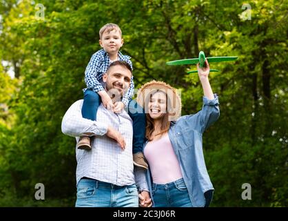 Netter Junge, der auf den Schultern seines Vaters sitzt und Mama, die Spielzeug Flugzeug am Wald hält Stockfoto