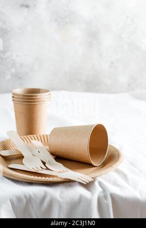 Leere Einweg-Öko-Utensilien - Teller, Tassen und Gabeln aus Holz und Papier maretials auf einer weißen Tischdecke gegen graue Wand backgroun Stockfoto