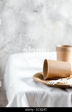 Leere Einweg-Öko-Utensilien - Teller, Tassen und Gabeln aus Holz und Papier maretials auf einer weißen Tischdecke gegen graue Wand backgroun Stockfoto