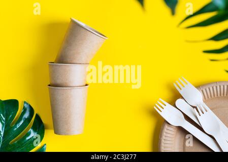 Leere Einweg-Öko-Utensilien - Teller, Tassen und Gabeln aus Holz und Papier auf gelbem Hintergrund mit tropisch-grünen Blatt, Kopierraum. Stockfoto